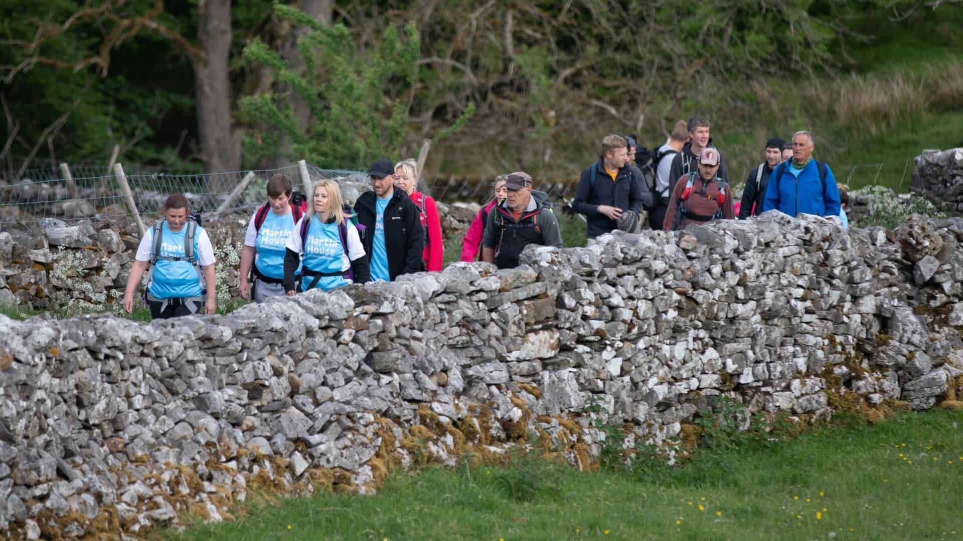 Large group of people walking on a path next to a stone wall.