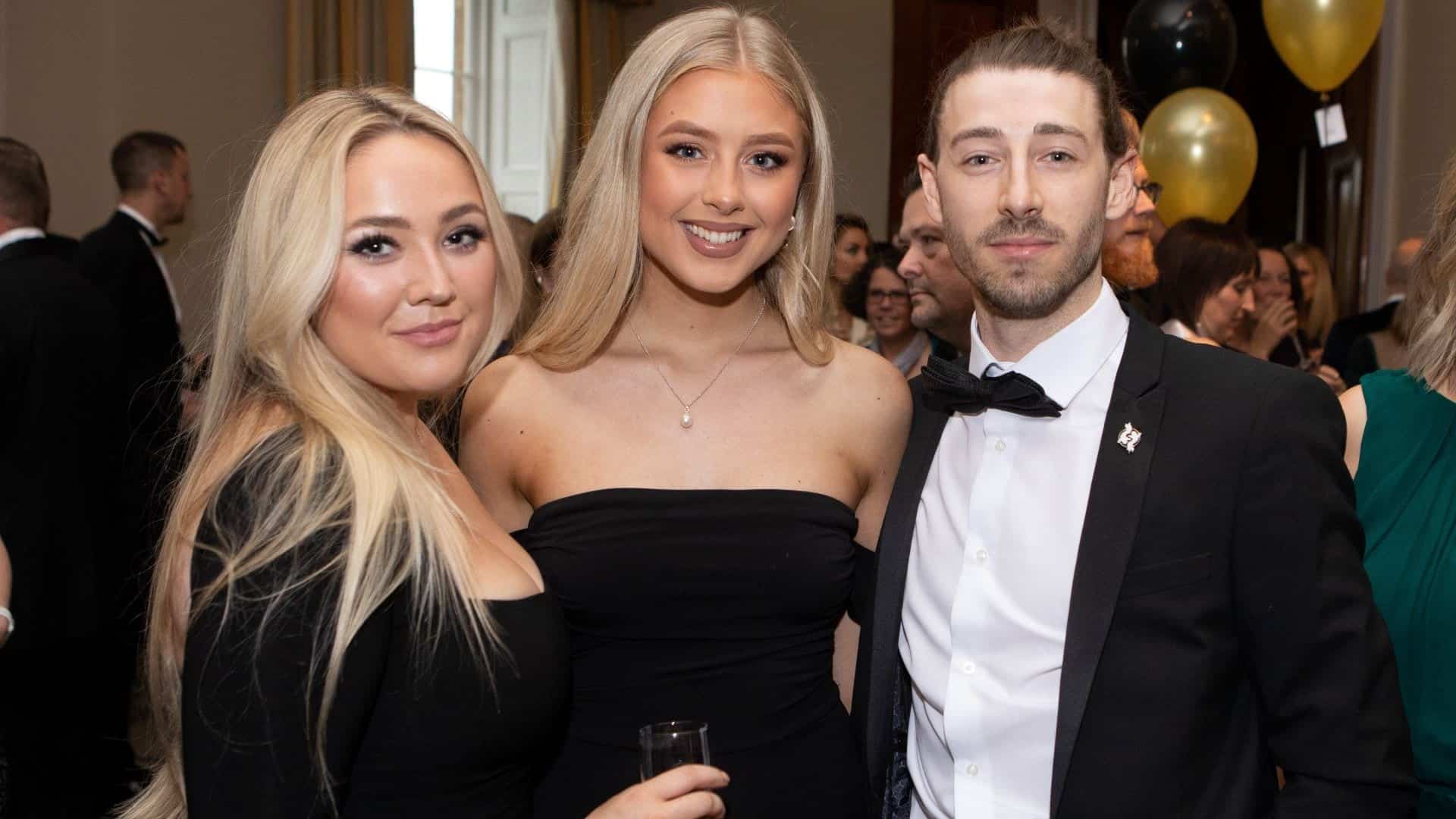 Three people, two women wearing ball gowns and one man wearing a suit.