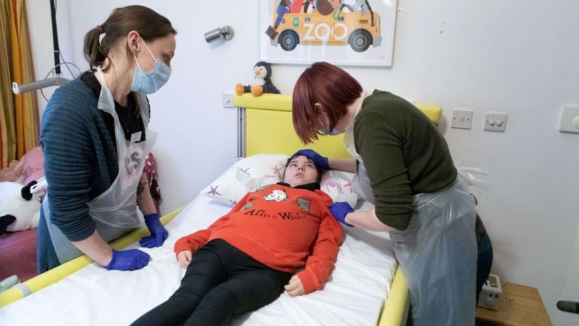 Two women caring for girl who is lying in a bed.