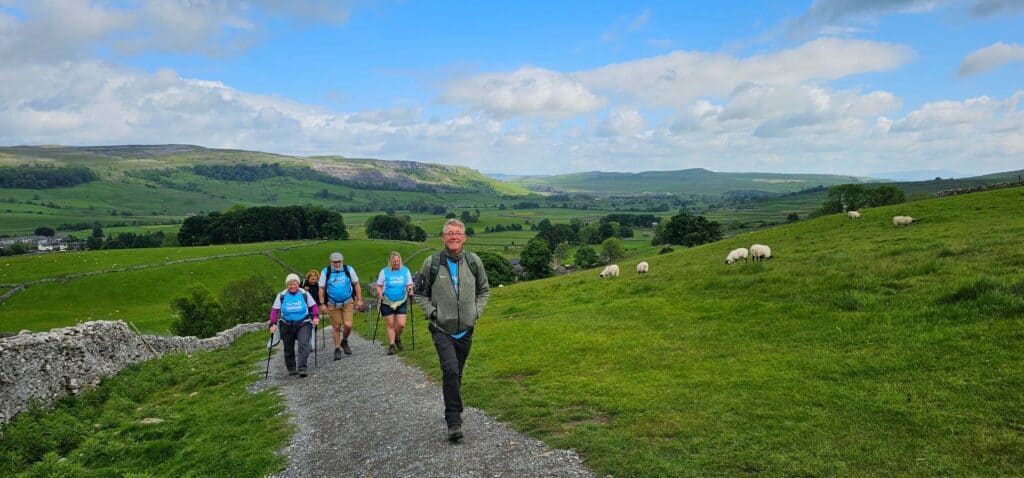 A group of smiling walkers in outdoor gear hike up a gravel path through rolling green hills in the Yorkshire countryside. Some are wearing light blue “Martin House” T-shirts and carrying walking poles. In the distance, sheep graze on grassy slopes under a bright blue sky with scattered clouds. Stone walls and patchwork fields stretch out across the landscape behind them.