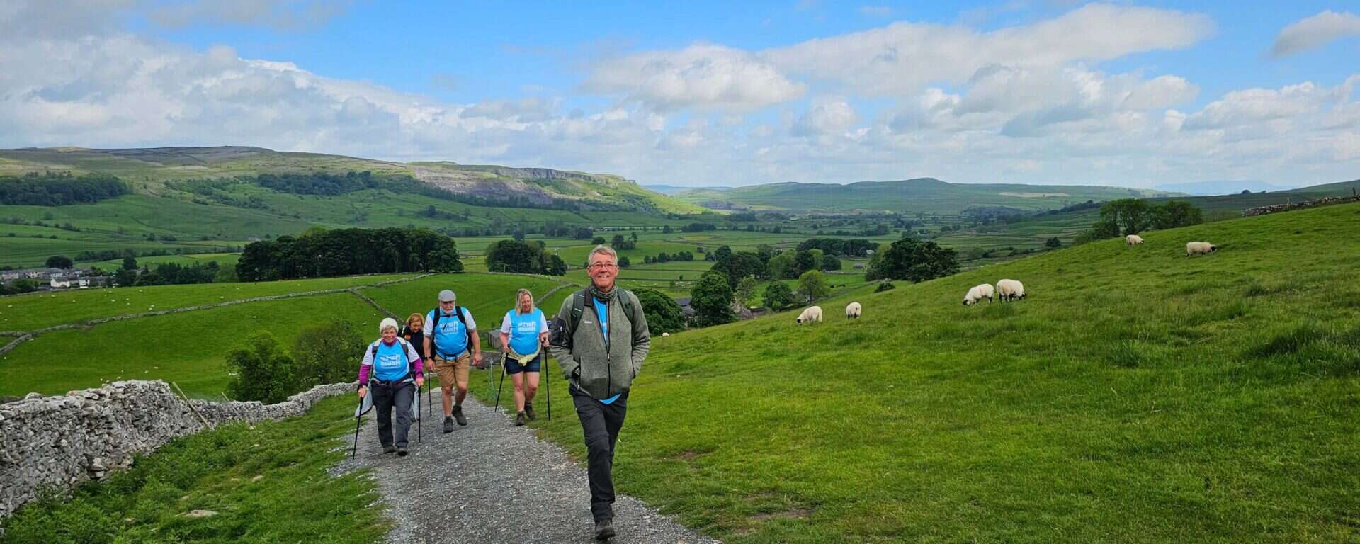 A group of smiling walkers in outdoor gear hike up a gravel path through rolling green hills in the Yorkshire countryside. Some are wearing light blue “Martin House” T-shirts and carrying walking poles. In the distance, sheep graze on grassy slopes under a bright blue sky with scattered clouds. Stone walls and patchwork fields stretch out across the landscape behind them.