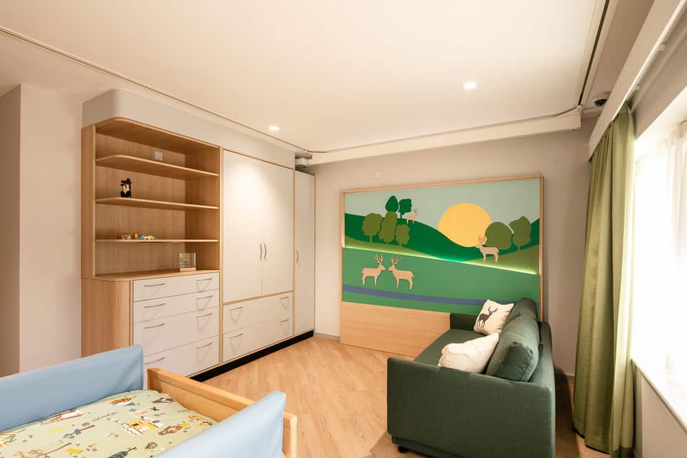 A bright and welcoming children's bedroom featuring wooden furniture, a green sofa, and a cheerful wall mural of deer and a sunrise in a green landscape. The room has warm lighting, a large wardrobe, open shelving, and a window with green curtains.