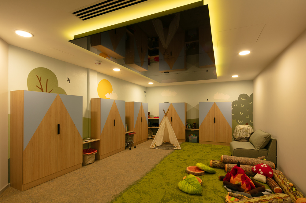 A cosy playroom with wooden cabinets decorated with mountain-shaped designs, a teepee tent in the center, and playful woodland-themed cushions and toys scattered on a green carpet. The walls are painted with trees, clouds, and a sun, creating a friendly, nature-inspired environment.