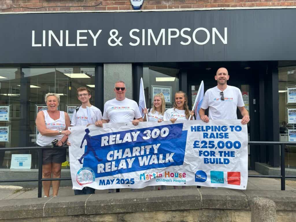 A group of six people stand smiling in front of a Linley & Simpson storefront, holding a banner that reads "RELAY 300 CHARITY RELAY WALK MAY 2025" and "HELPING TO RAISE £25,000 FOR THE BUILD." They are wearing white Martin House Children's Hospice t-shirts. The banner includes logos for Martin House, Dale Eddison, Linley & Simpson, and Hardisty.
