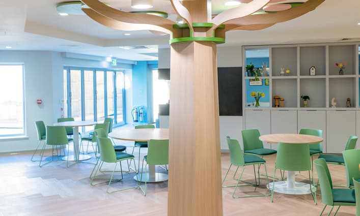A modern and inviting dining area with round tables and light green chairs, centered around a large wooden pillar designed to look like a stylized tree. The room is well-lit with natural light from tall windows and has light wooden flooring and built-in shelving with decorative items.