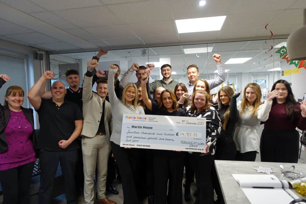 A large group of colleagues stand together in an office, smiling and raising their arms in celebration while holding a giant cheque for £14,917.75 made out to Martin House Children’s Hospice, donated by Training at Work Group.