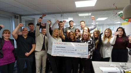 A large group of colleagues stand together in an office, smiling and raising their arms in celebration while holding a giant cheque for £14,917.75 made out to Martin House Children’s Hospice, donated by Training at Work Group.